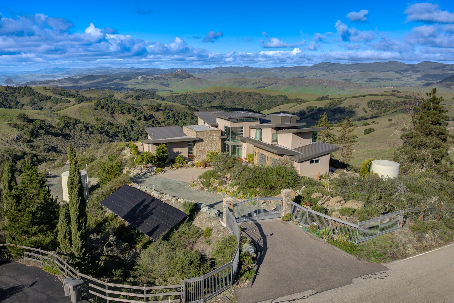 Drone view of a contemporary hillside home, gated driveway, and surrounding green hills beneath a blue sky.