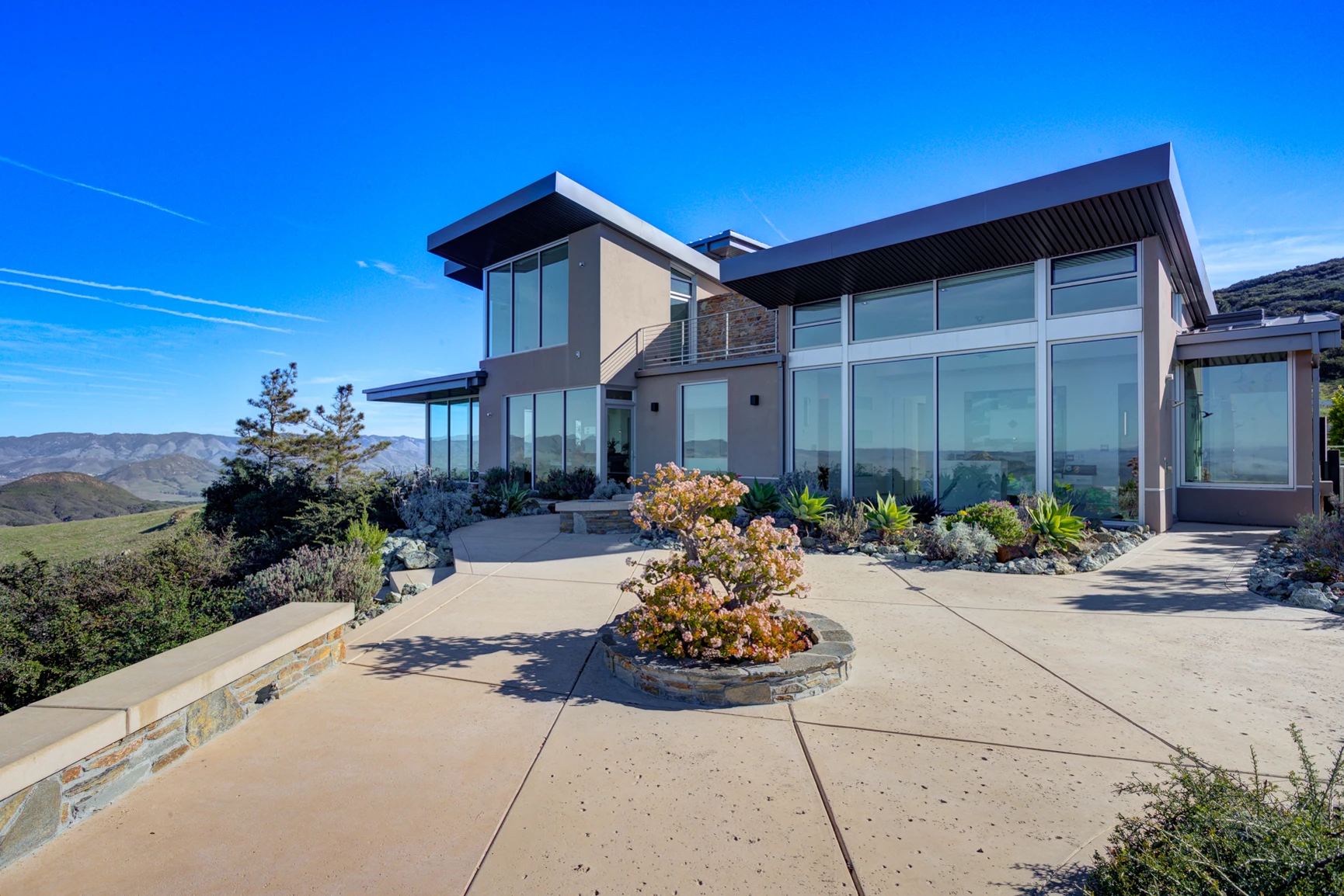 Contemporary hillside home with angular rooflines, tall windows, stone accents, and a landscaped terrace beneath a blue sky.
