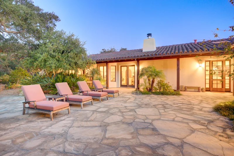 Stone terrace and loungers outside an illuminated home at dusk.